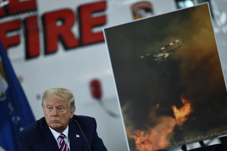 Trump está sentado em uma reunião sobre incêndios florestais, com uma imagem de um helicóptero jogando água em um incêndio ao fundo.