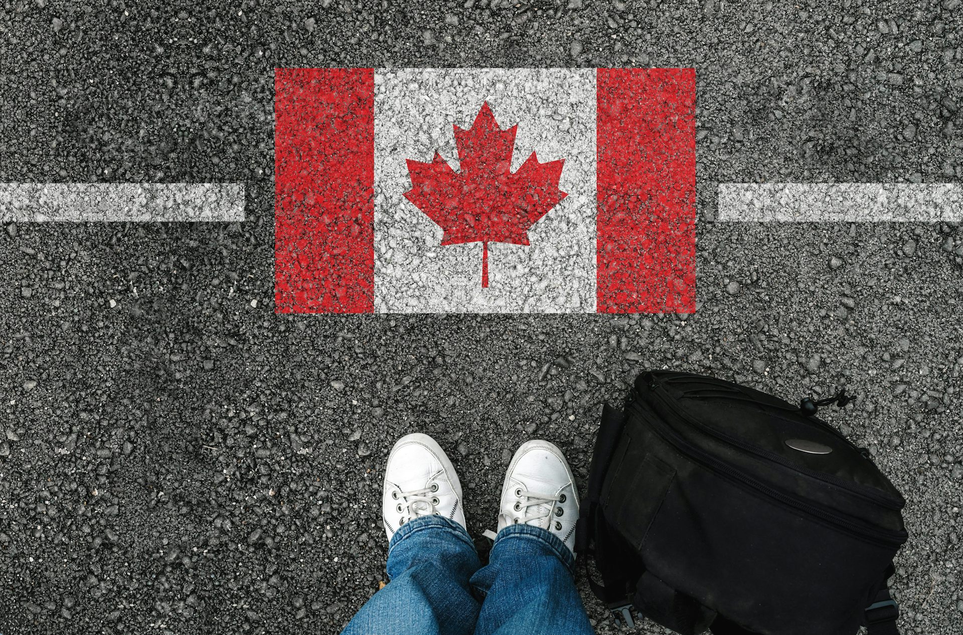 A person's feet and back back are seen standing near a Canadian flag painted on asphalt