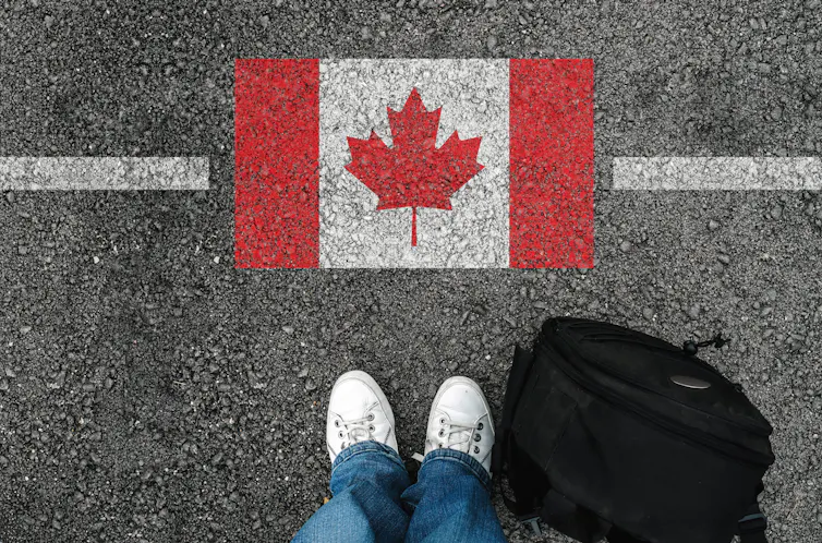 A person's feet and back back are seen standing near a Canadian flag painted on asphalt