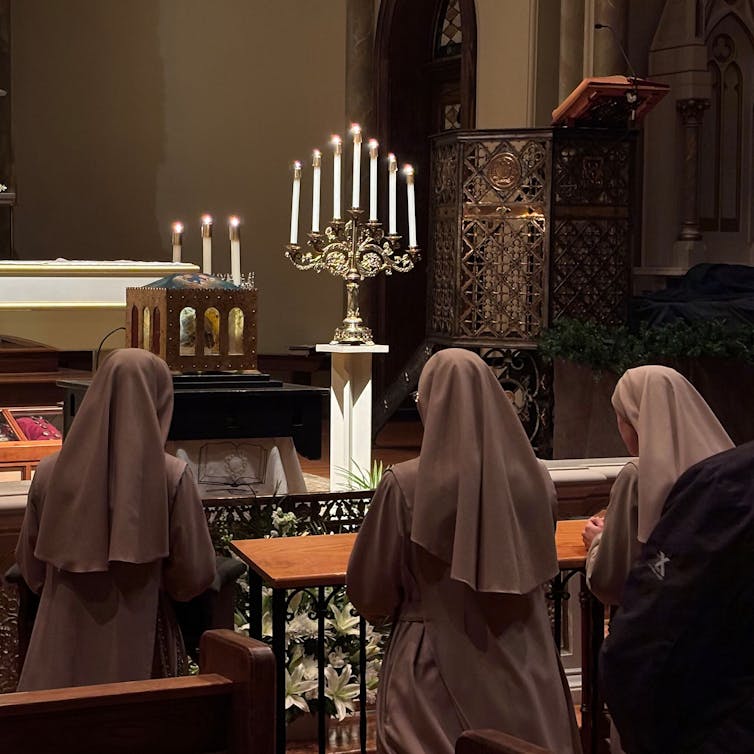 Three women in nuns' habits, seen from the back, kneel at the front of a church, looking toward a small box on a pedestal.