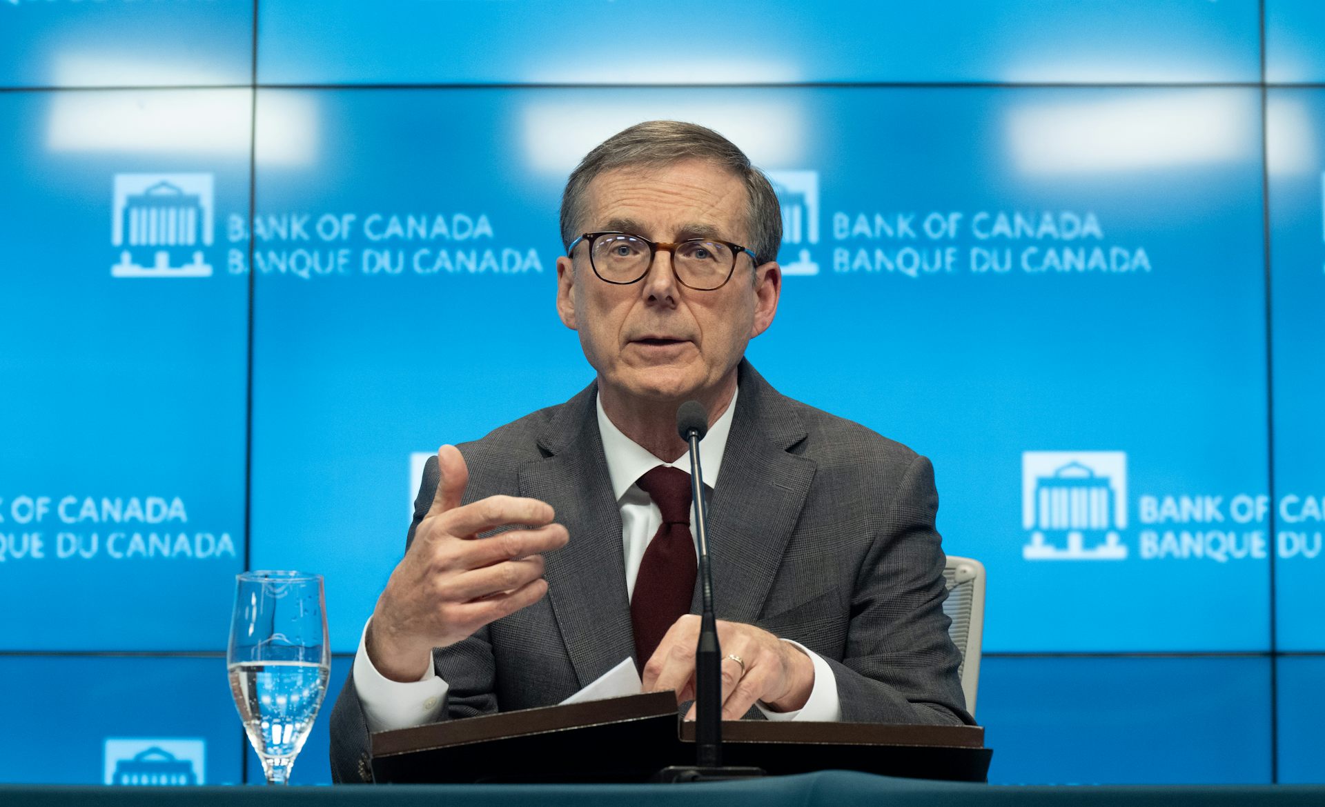 A man with grey hair and glasses speaks into a microphone from behind a conference table. Behind him is a blue backdrop that says 'Bank of Canada'
