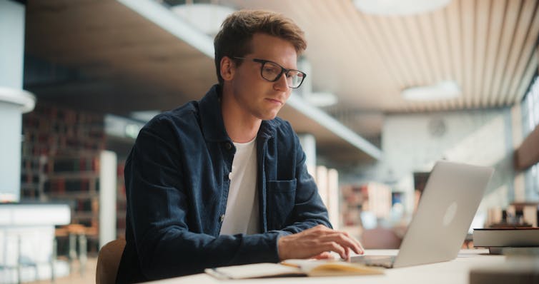 A young man wearing glasses sits at a desk using a laptop.
