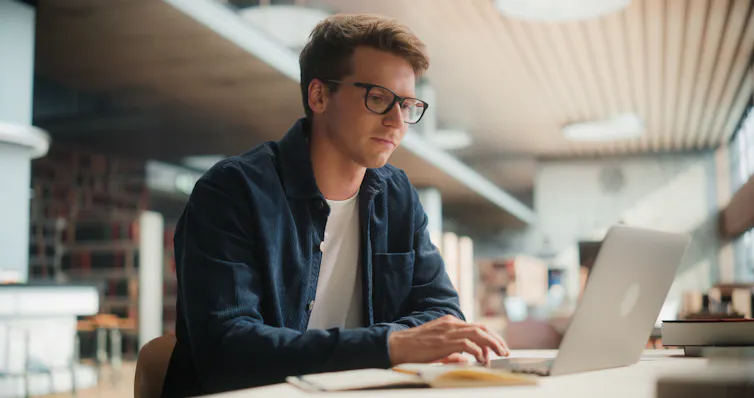 A young man wearing glasses sits at a desk using a laptop.