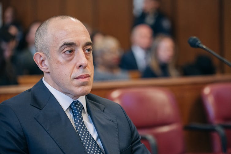A mostly bald man in a suit and tie sitting in a large room.