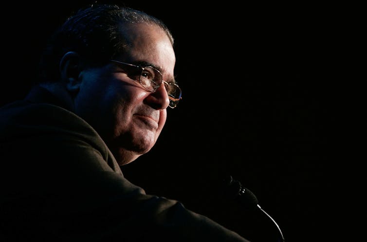 A man with glasses and dark hair standing at a microphone.