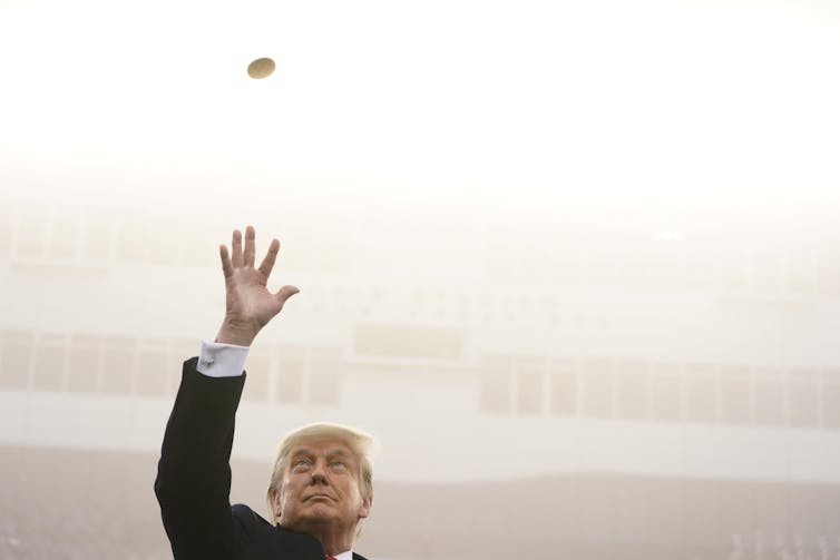 A man with fluffy blond-grey hair tosses a coin in the air.