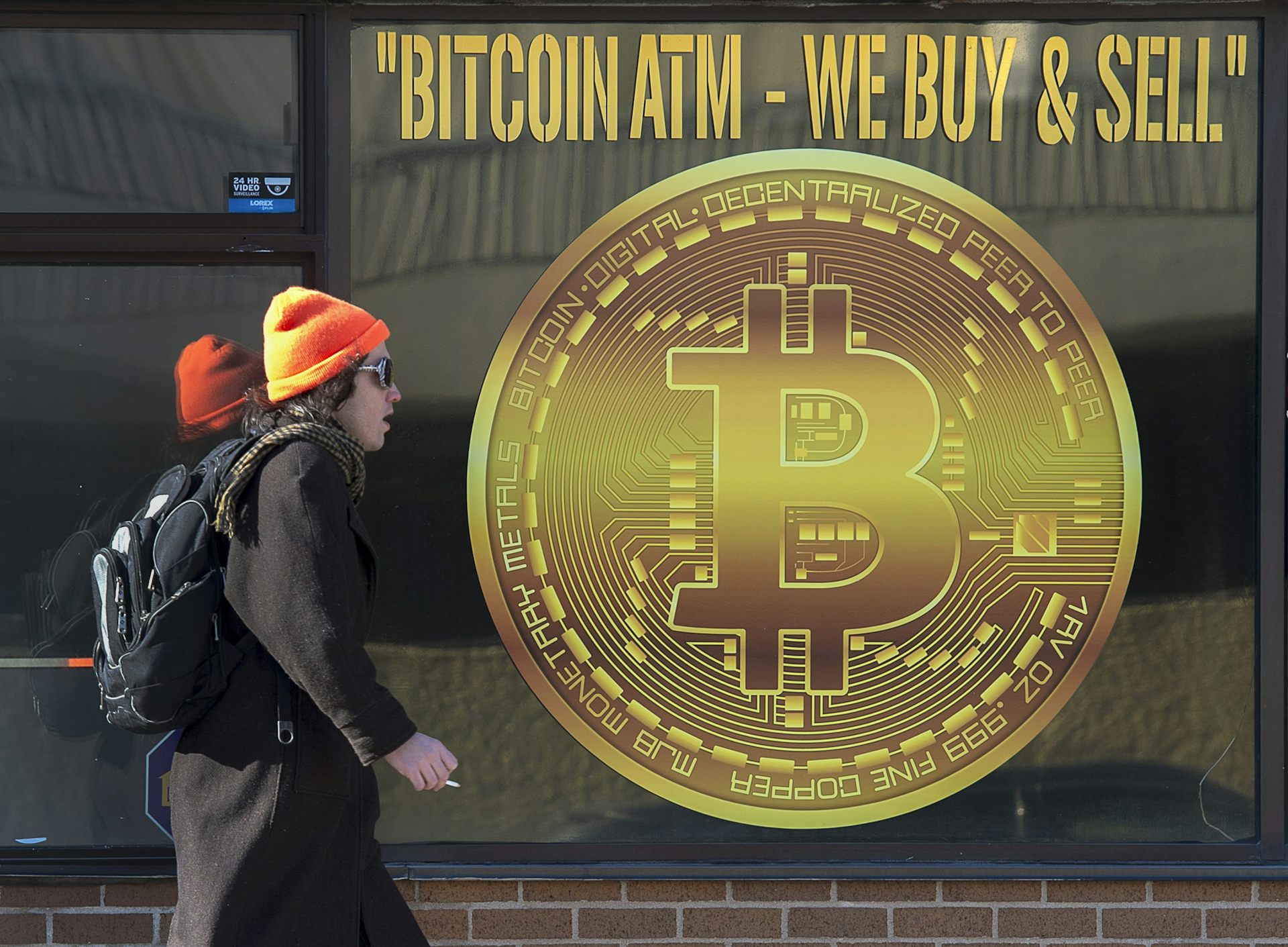 A woman in a dark coat wearing an orange toque walks past a sign advertising Bitcoin.