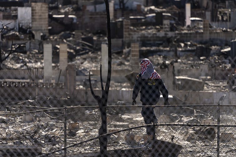 a person in uniform and an american flag wrapped around their head walks through a burned-out area