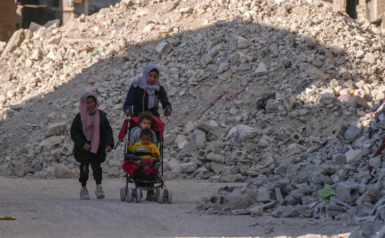 A woman and tow children walk past rubble.
