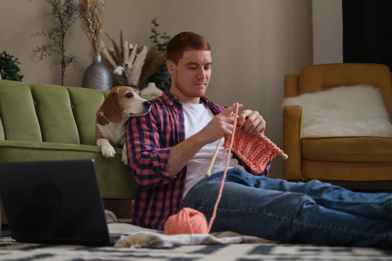 Um homem tricota sentado no chão. Seu beagle está sentado no sofá atrás dele.