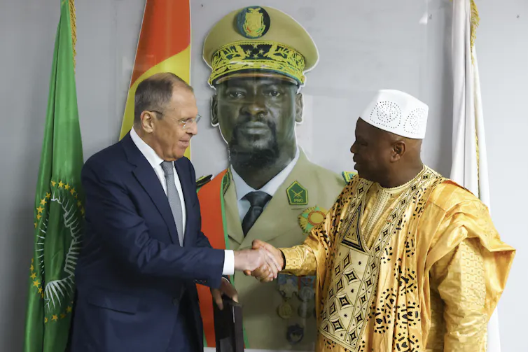 A Black man in a west african outfit shakes hands with a white man in a dark blue suit.