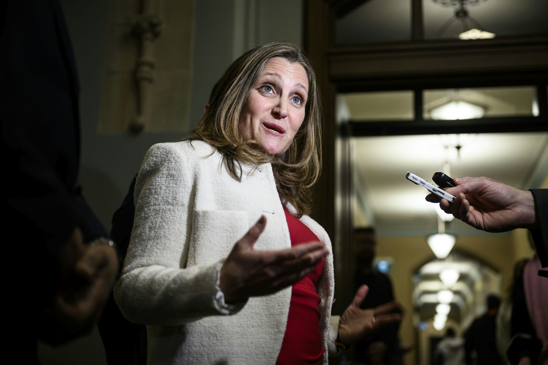 A woman with dark blond hair in a white jacket over a red blouse speaks to reporters.