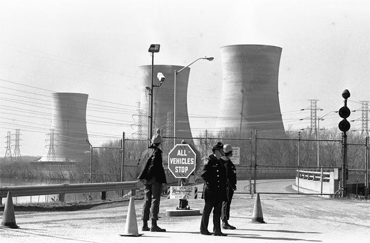 a black and white photograph of three men standing in the foreground near a stop sign while there are three cooling towers visible in the background