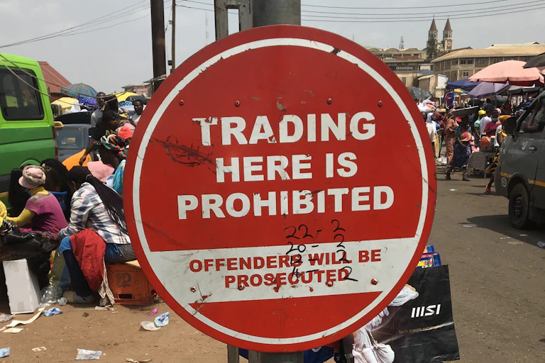 A sign on a pole saying Trading Here is Prohibited, with a busy street scene in the background