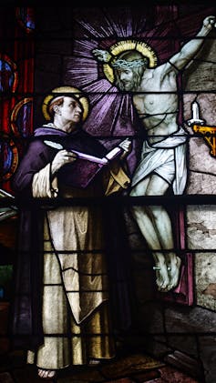 A man in a white and purple robe, with a halo around his head, stands close to a crucified Christ, who seems to speak to him.