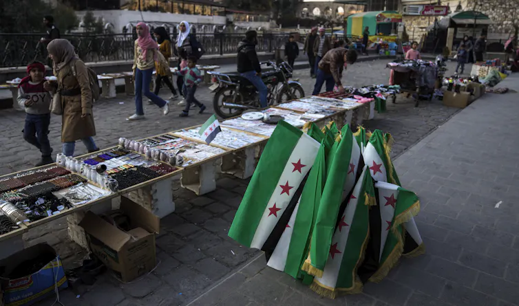 People walk past street vendors and a collection of Syrian flags