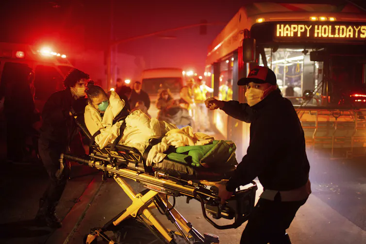 Emergency responders push a person laying on an ambulance stretcher with smoky air and a city bus nearby.