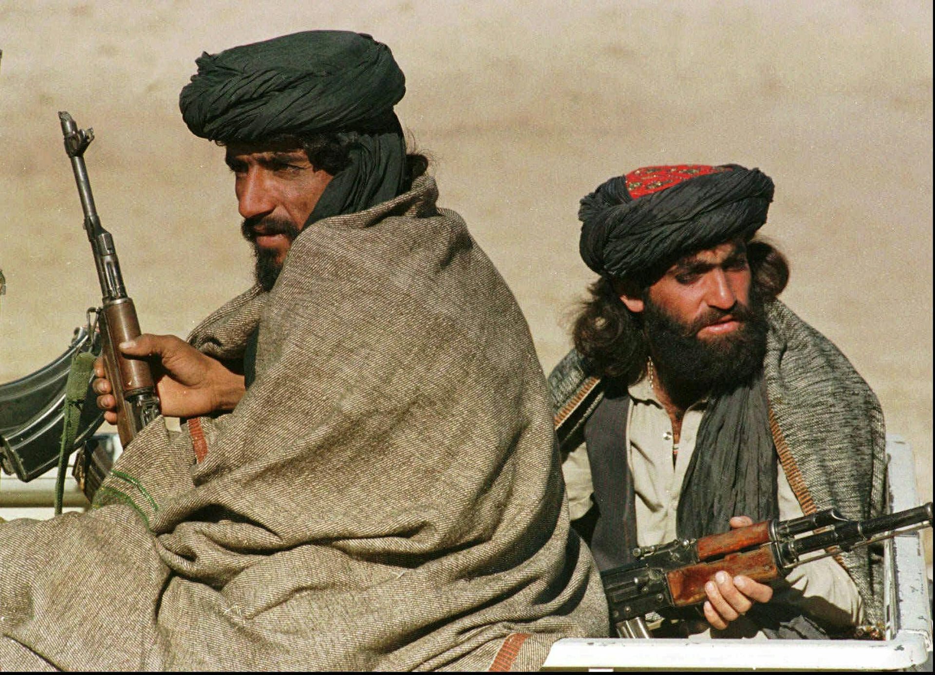 Two men with thick beards hold weapons as they sit in the back of a vehicle.