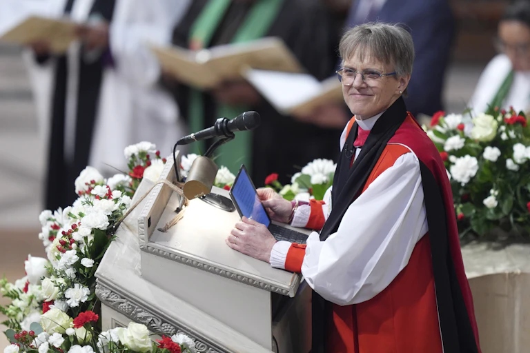 A gray-haired, spectacled woman in white and red priestly attire stands at a lectern adorned with flowers, holding a tablet and smiling as she speaks.