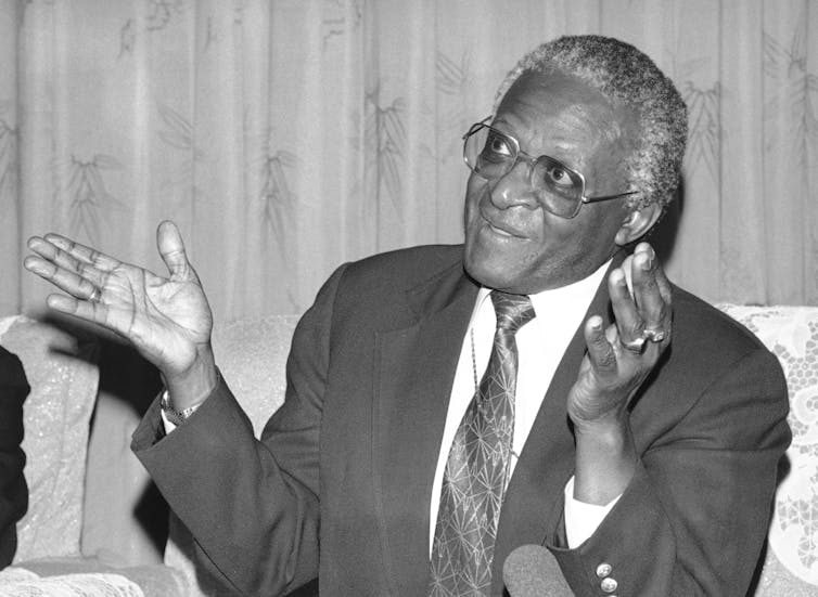 A black-and-white photo shows a grey-haired, spectacled man in a suit and tie, speaking and gesturing with his hands.