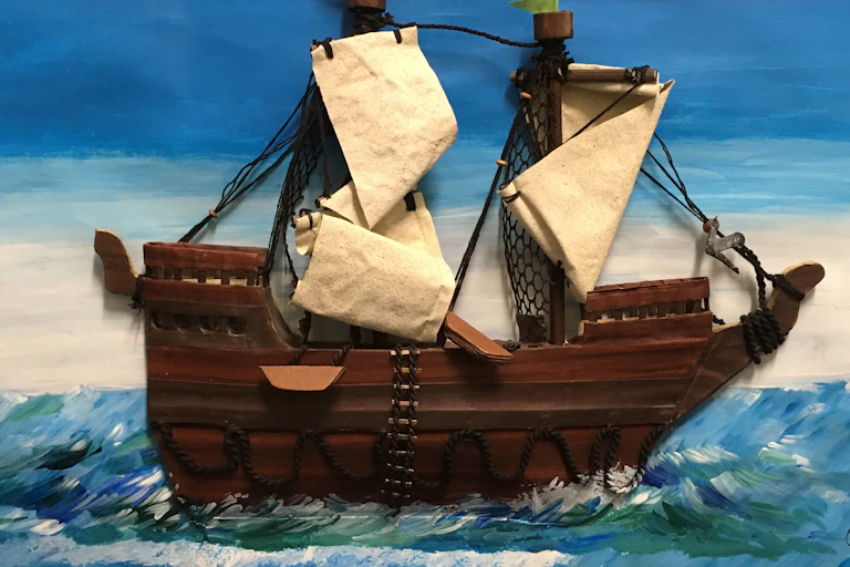 A sailing ship made out of found objects is seen in a cardboard frame.