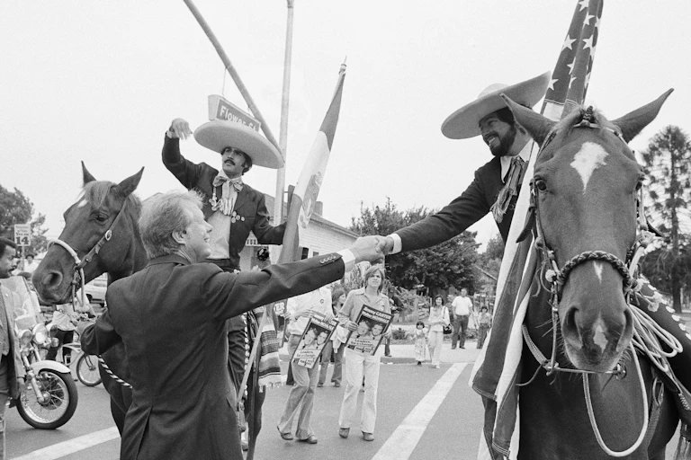 Jimmy Carter shakes the hand of a man riding a horse while wearing a sombrero and carrying an American flag.