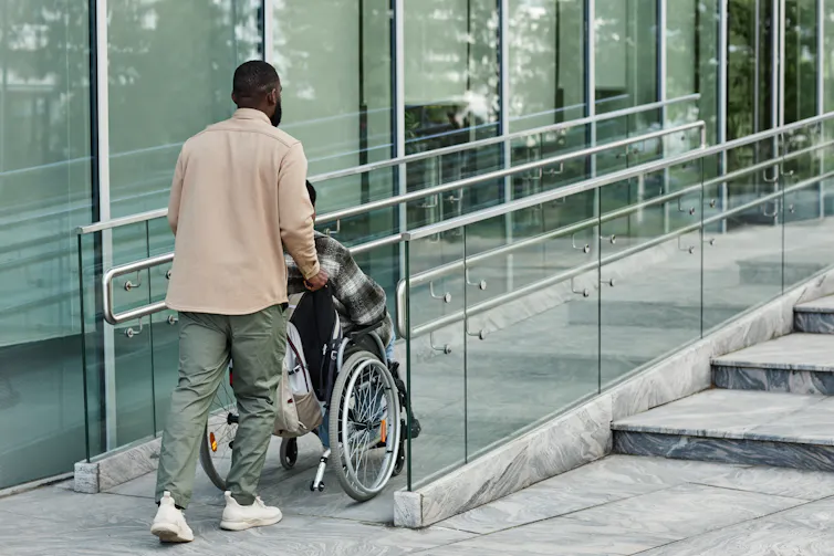 A person pushes someone in wheelchair up a ramp.