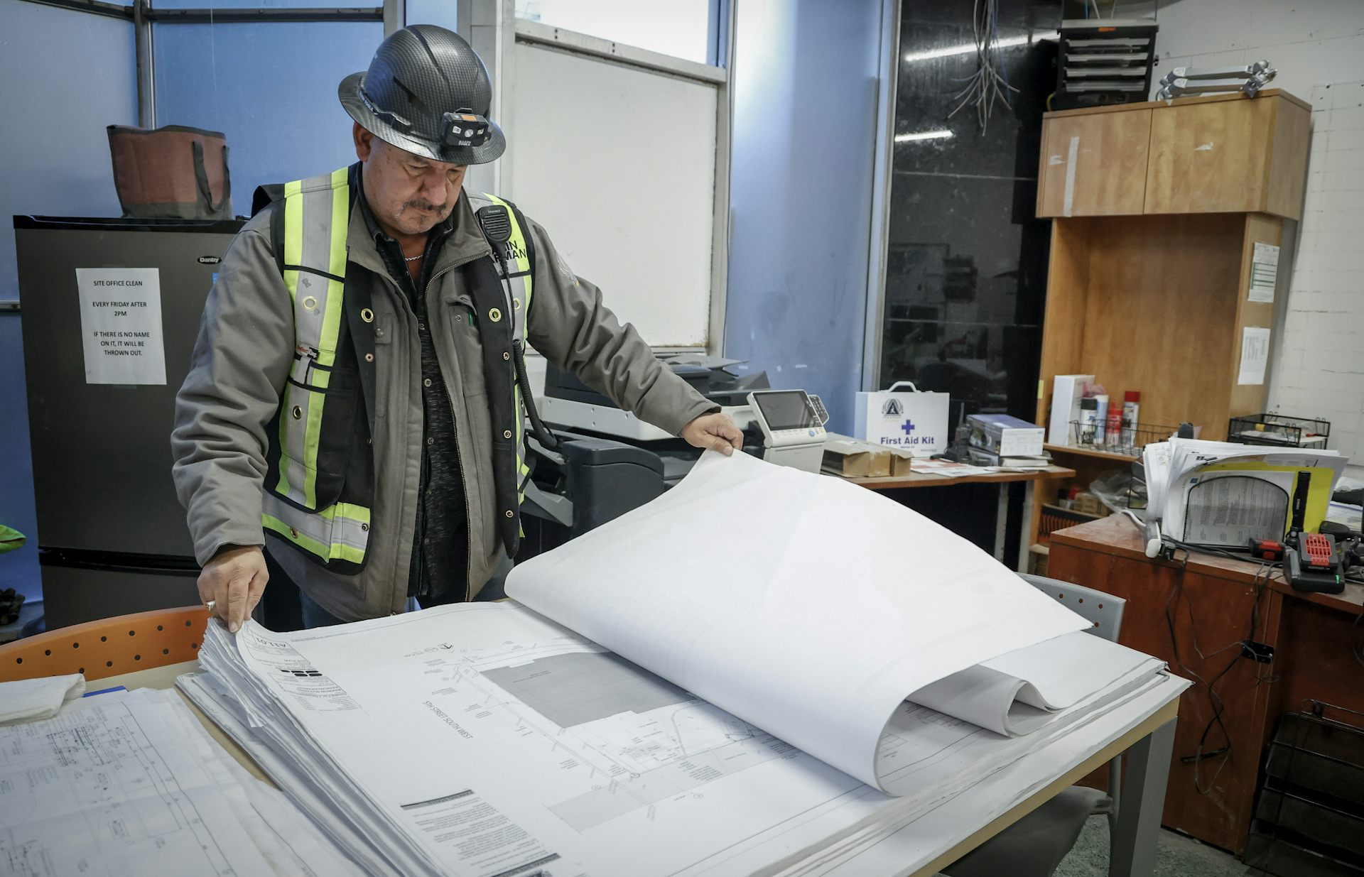 Un hombre que lleva una chaqueta Hardhat y Hi-Vision mirando el draft en la oficina