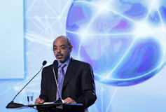 A man in a dark suit and lilac tie speaks into a microphone.