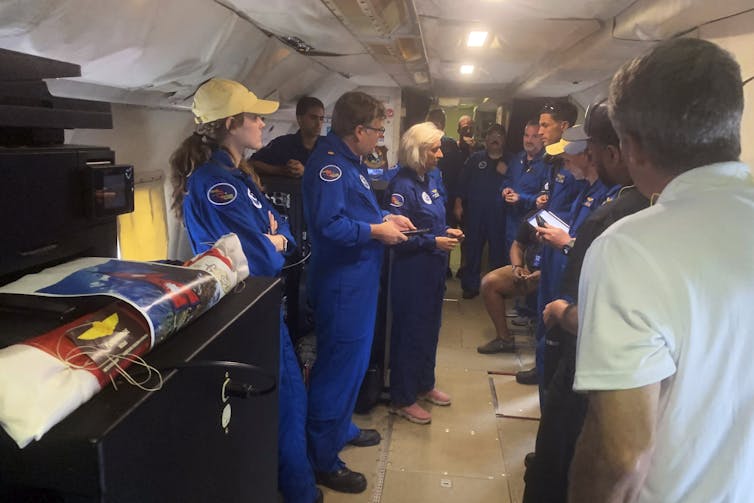a group of people wearing identical blue suits and some wearing hats stand and talk in the hull of a plane