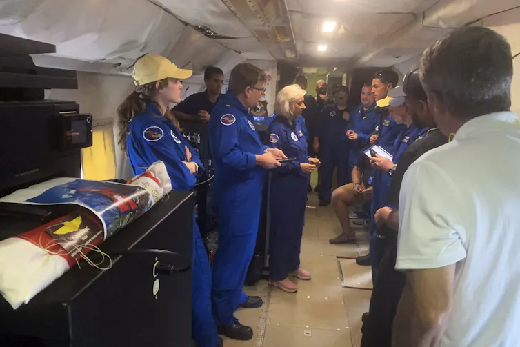 a group of people wearing identical blue suits and some wearing hats stand and talk in the hull of a plane