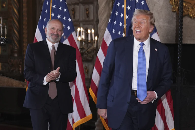 A bearded man in a dark suit and tie clasps his hands and smiles as another man in a dark suit and tie appears to move next to him, with American flags in the background