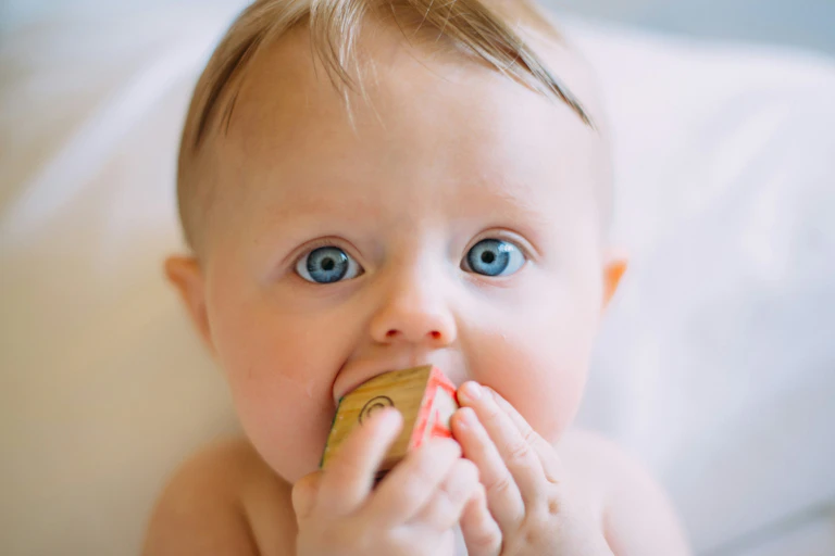 Baby with wide, blue eyes and blonde hair chewing a wooden cube.