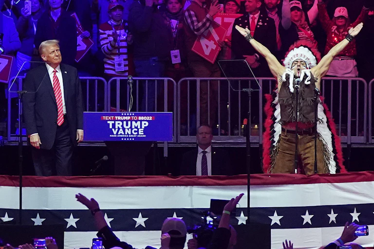 Weird-looking orange-faced man with an extra long tie looking smugly on at man dressed in native Indian dress doing the actions to YMCA.