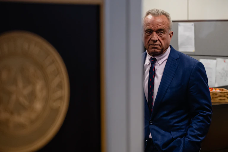A pensive man in a blue jacket, white shirt and dark tie, standing near a doorway.