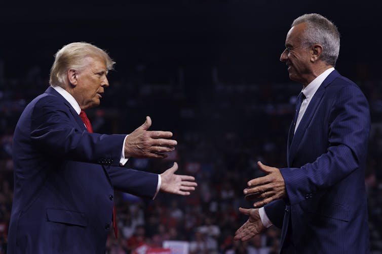 Two men in blue suits greet each other with open arms.