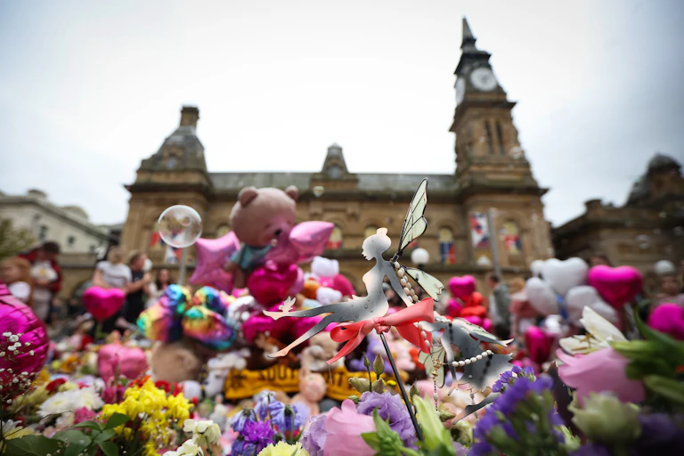 Close up of flowers and gifts, many of which are pink, at a vigil for the Southport victims, with a building, the Atkinson cultural centre, visible in the background"