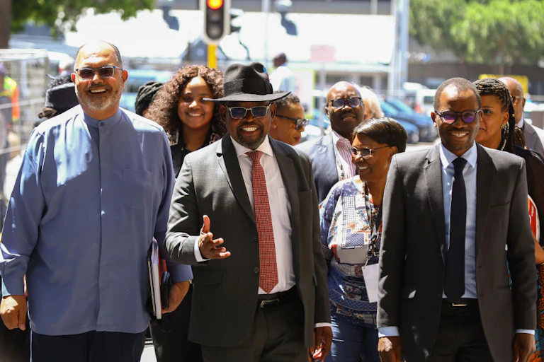 Smiling men and women walk towards the camera outdoors. One man in suit and tie is wearing a hat.