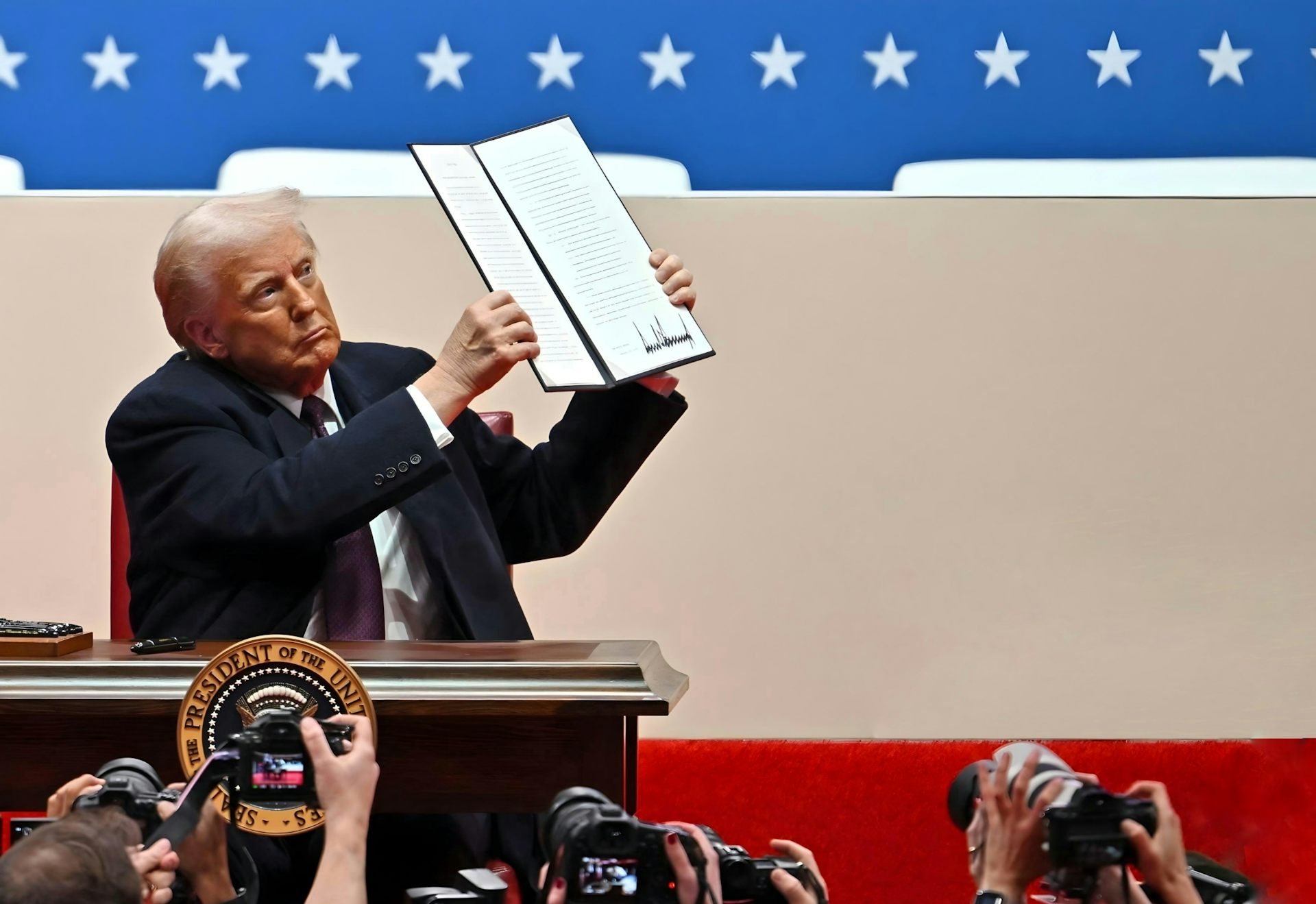 Donald Trump holds up a text that includes what appears to be his signature in front of people with cameras.