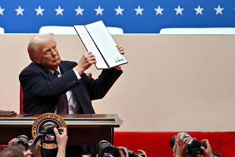 Donald Trump holds up a text that includes what appears to be his signature in front of people with cameras.
