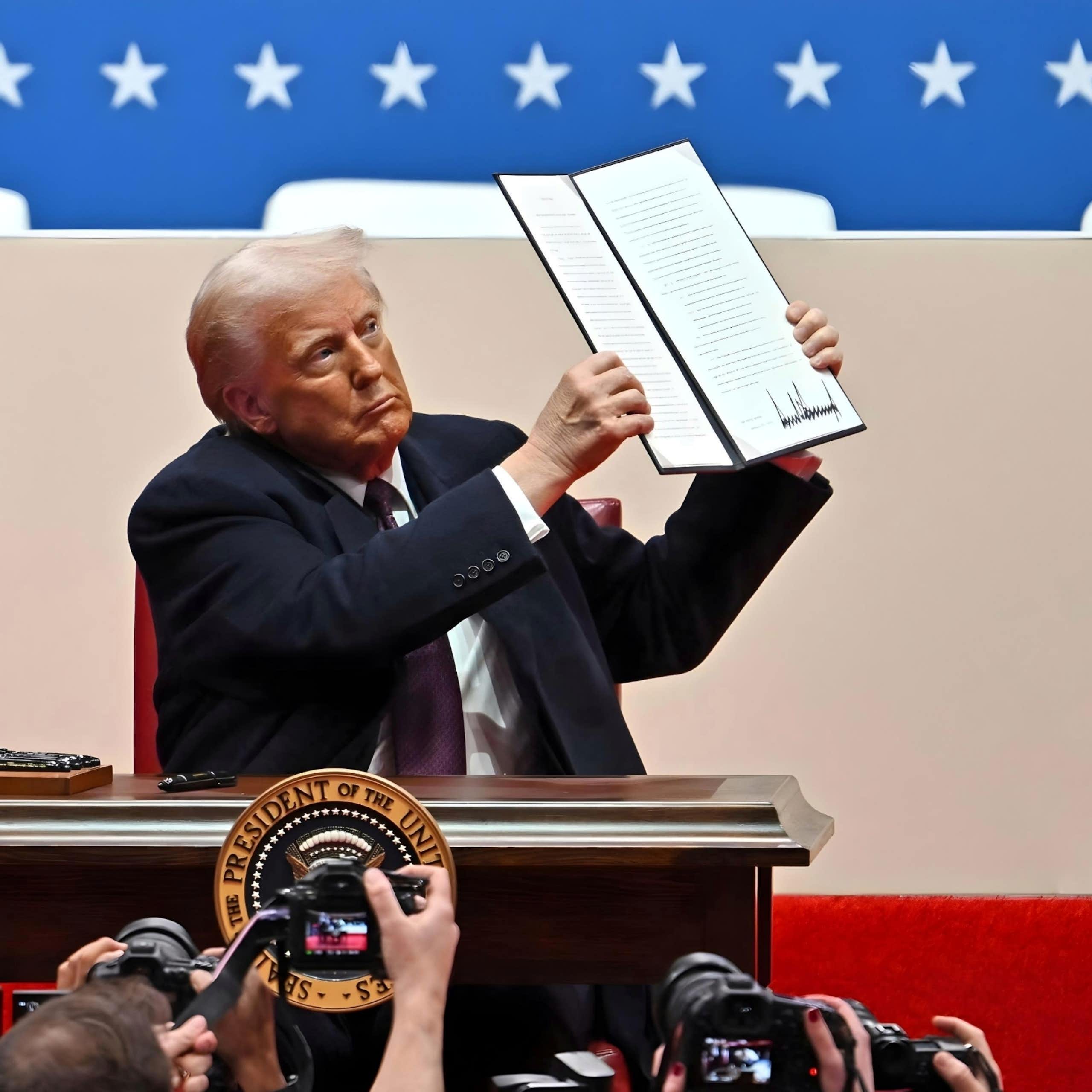 Donald Trump holds up a text that includes what appears to be his signature in front of people with cameras.