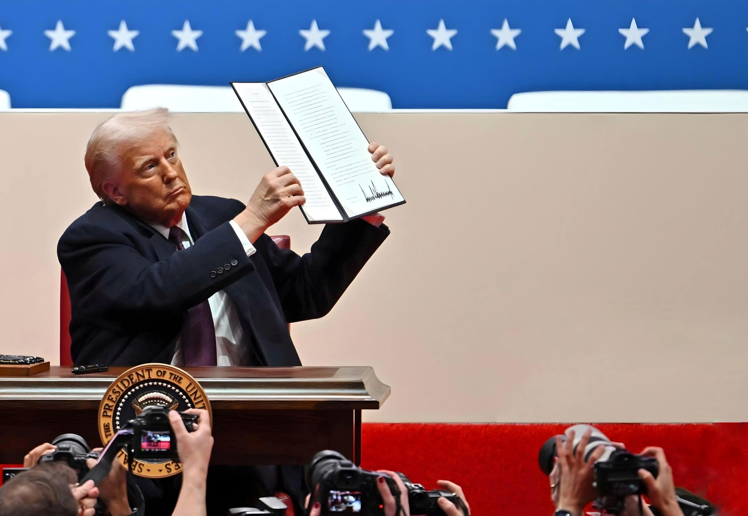 Donald Trump holds up a text that includes what appears to be his signature in front of people with cameras.