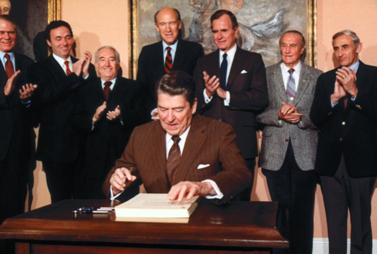 President Reagan signs a document while surrounded by men in suits.
