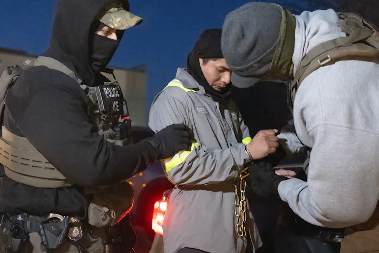Two men are seen holding a man with a grey sweater and his hands in handcuffs.