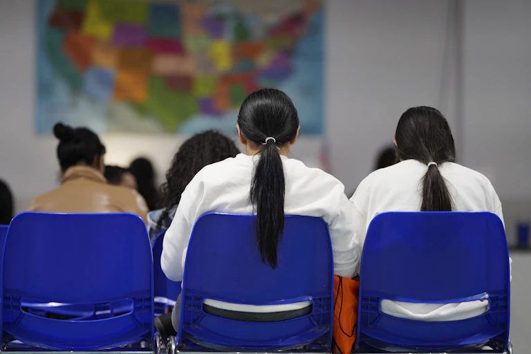 Women with dark hair sit in blue seats and are seen from behind facing a map of the United States.
