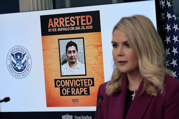 A blonde woman wearing a maroon jacket stands in front of a screen that shows a photo of a man with the words 'Arrested, convicted of rape' around the photo.