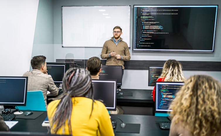 a teacher speaks to students in a classroom with a large screen displaying computer code