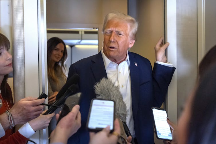 A sleepy-looking elderly man with greyish-blond hair and facial bronzer takes questions from reporters.