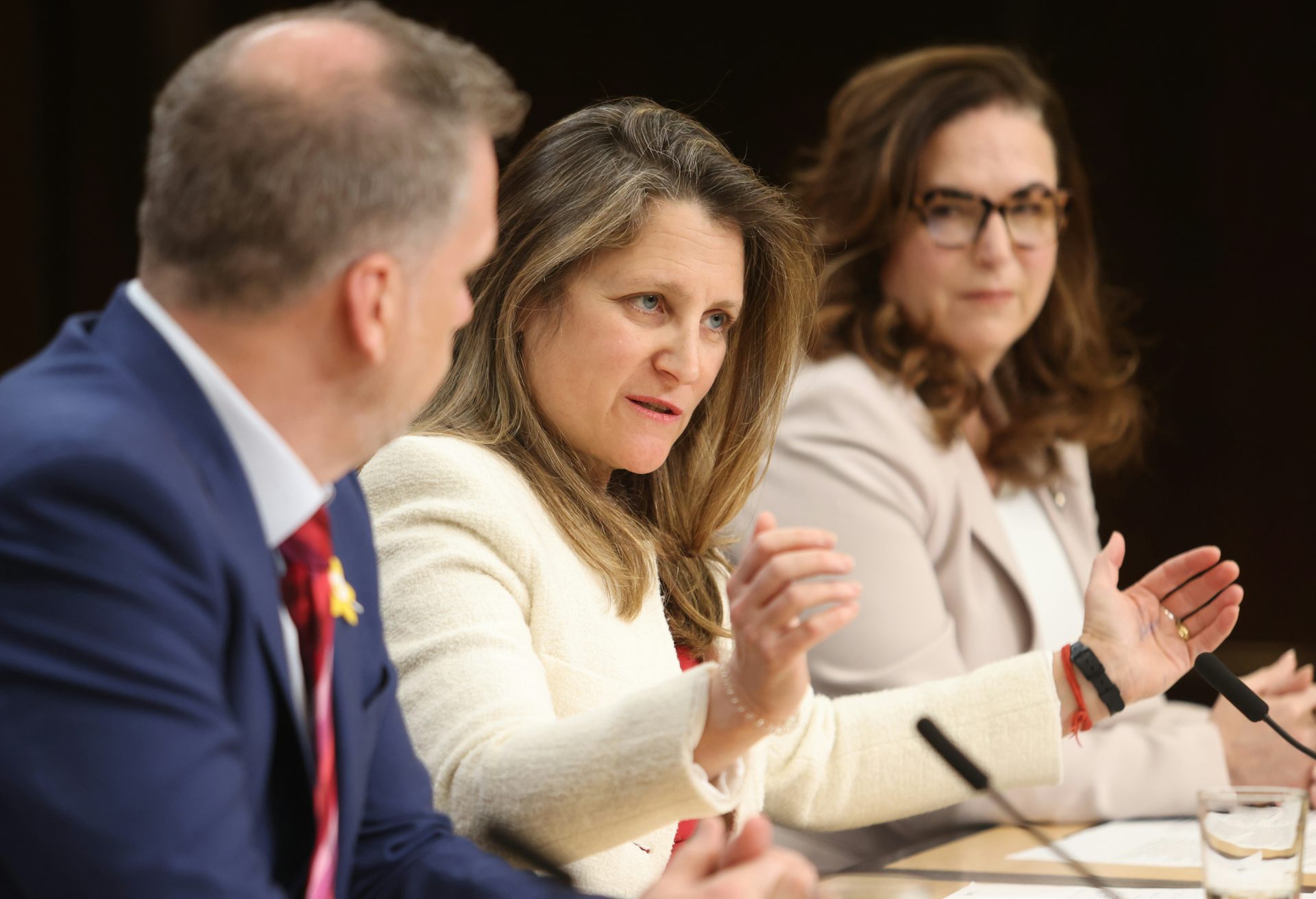 A woman speaking and gesting with her hands on a table with a microphone by a man and another woman