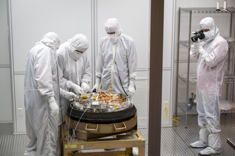 Four people in white, protective bunny suits unpacking a golden capsule.